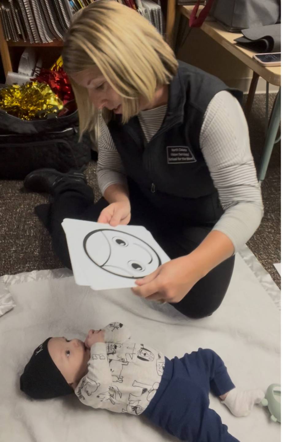 A woman sits on a floor holding an image of a face above an infant who is laying on the floor.